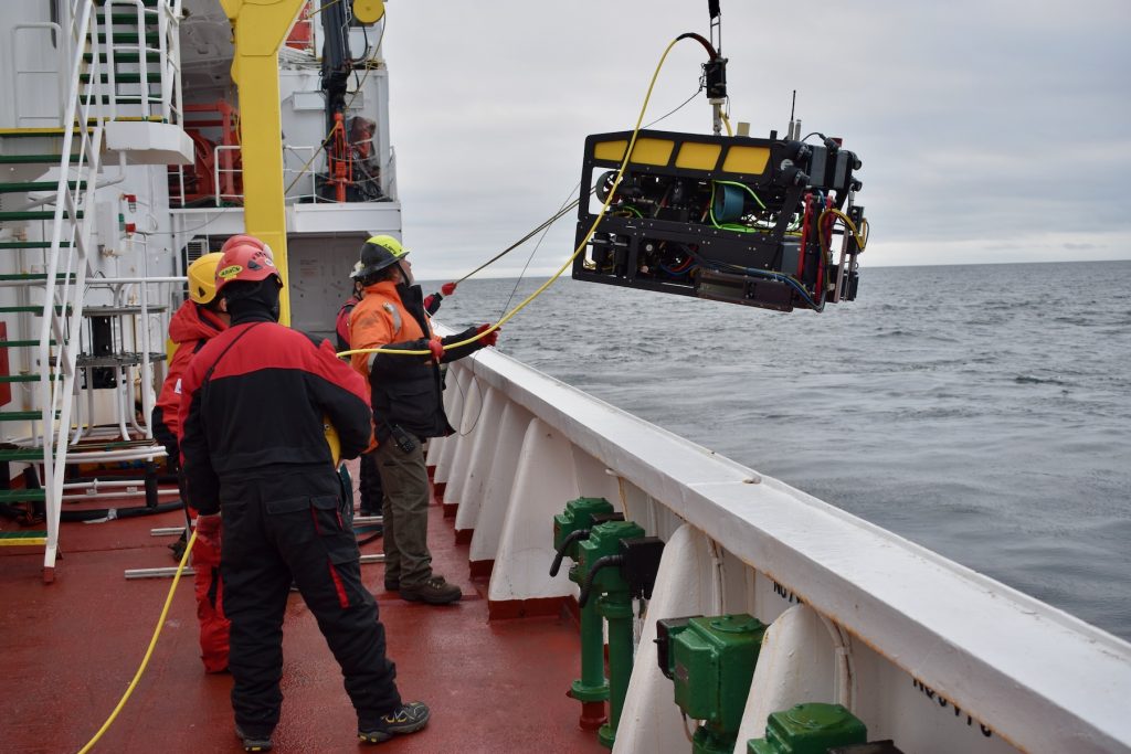 Marine operations crew members launch a robotic submersible over the side of a research ship into the water. The crew members are on the left side of the frame. The submersible has a yellow float, black frame, and a red mechanical arm, and is equipped with various scientific instruments. A black tether extends offscreen at the top of the frame to a crane on the research ship. Marine operations crew members are holding two yellow tethers, one to carry video from the submersible’s cameras and one to stabilize the submersible during deployment. The background is gray-blue ocean and overcast gray sky.