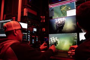 A pilot steers a remote deep-sea robot from the control room aboard a research ship. The pilot is wearing a white trucker hat and a gray hooded sweatshirt. He is seated in front of a wall of six video monitors displaying live camera feeds and data from the robot as it moves along the seafloor. He controls the robot with a joystick. In the background is a tower of computer equipment. The frame is illuminated with red light.