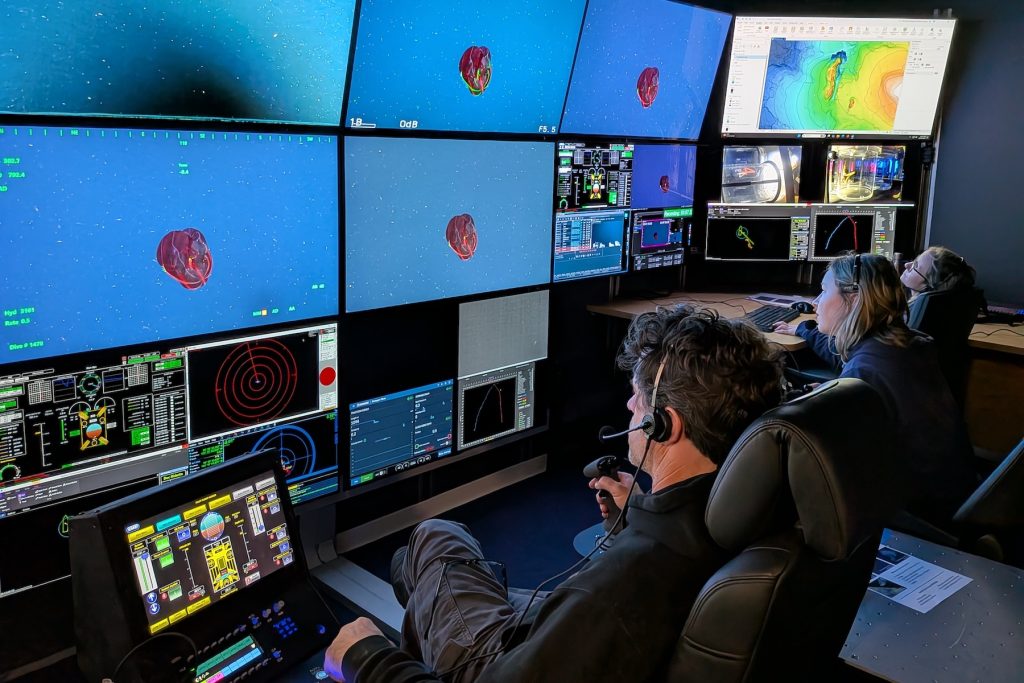 A submersible pilot and two biologists sit in black leather chairs and watch video monitors in the dark submersible control room aboard a research ship. The pilot is in the foreground. He has wavy salt-and-pepper hair, is wearing a headset, a black hooded sweatshirt, and gray pants, and is holding a black rubber joystick. The biologist in the center has short blonde hair. She is wearing a headset and a navy-blue jacket. The biologist in the background has short blonde hair. She is wearing glasses and a headset. All three are watching a wall of video monitors on the left side of the frame displaying a video feed and data from the deep sea. A bright red comb jelly swimming in open blue water is displayed on the two monitors at the center of the wall.
