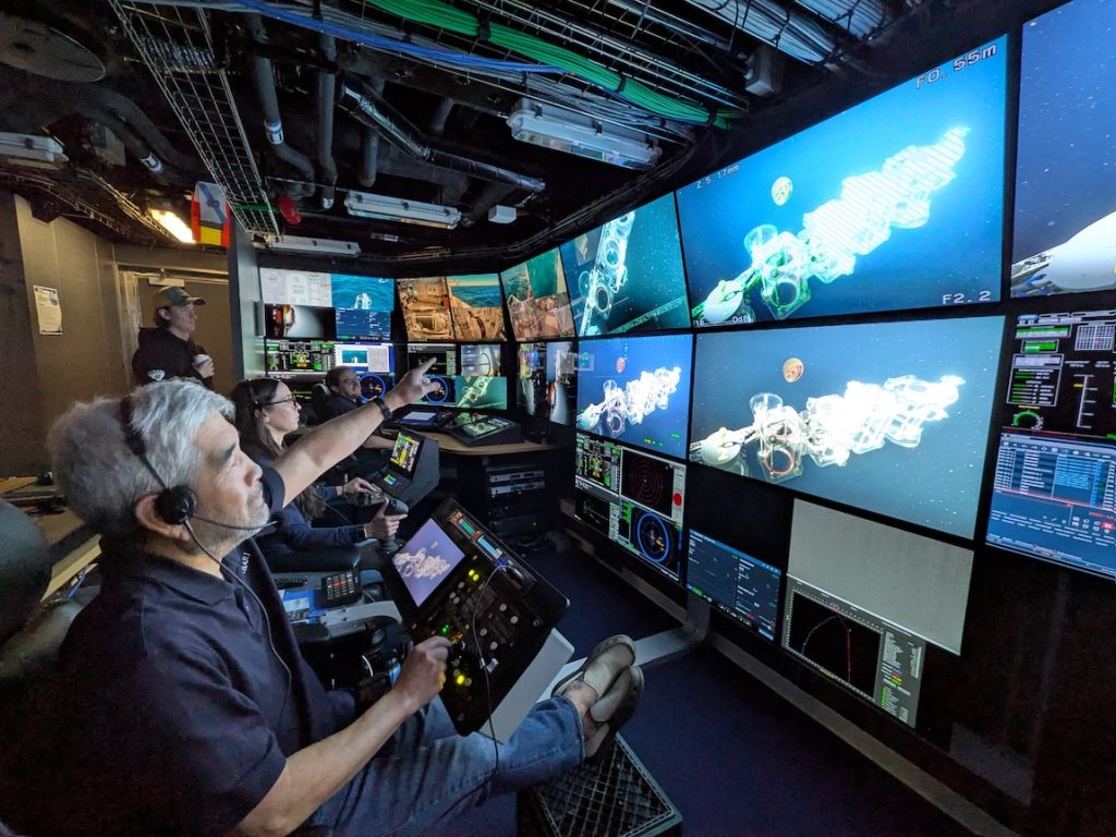 An MBARI researcher with short gray hair and a gray beard wearing a black headset, navy blue polo, blue jeans, and tan slip-on shoes points to a wall of video monitors in the dark submersible control room of a research ship. The researcher is seated in a black leather chair with his legs up on a black plastic milk crate. In the background are two submersible pilots seated in chairs and operating submersible controls and a third submersible pilot holding a cup of coffee. The video monitors on the right side of the frame are displaying a live feed from a deep-sea submersible showing an array of clear plastic sampling canisters and a bright red jelly.