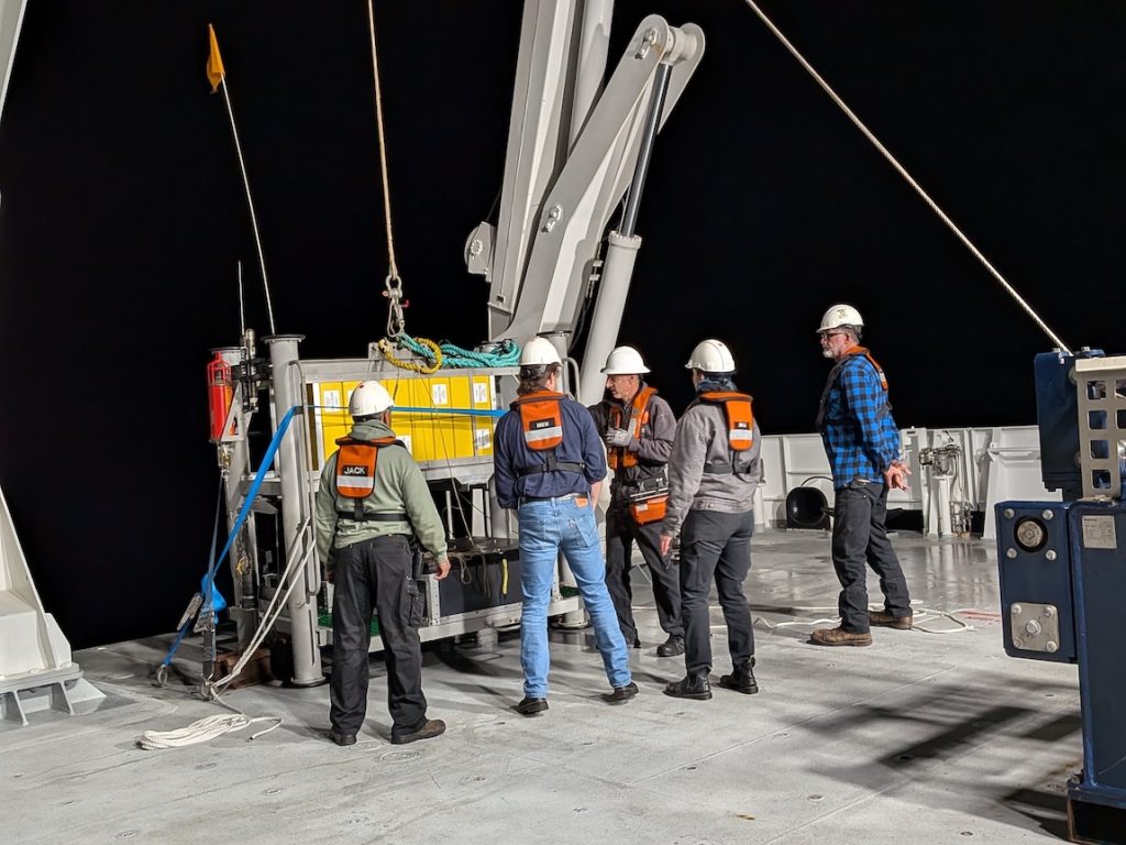 Five members of MBARI’s marine operations crew discuss deployment of a scientific instrument from the gray metal deck of a research ship. The crew members are wearing white hard hats and orange life vests over their clothes. The instrument on deck has yellow floats, a silver metal frame, and a black plastic trunk. The background is black night sky.