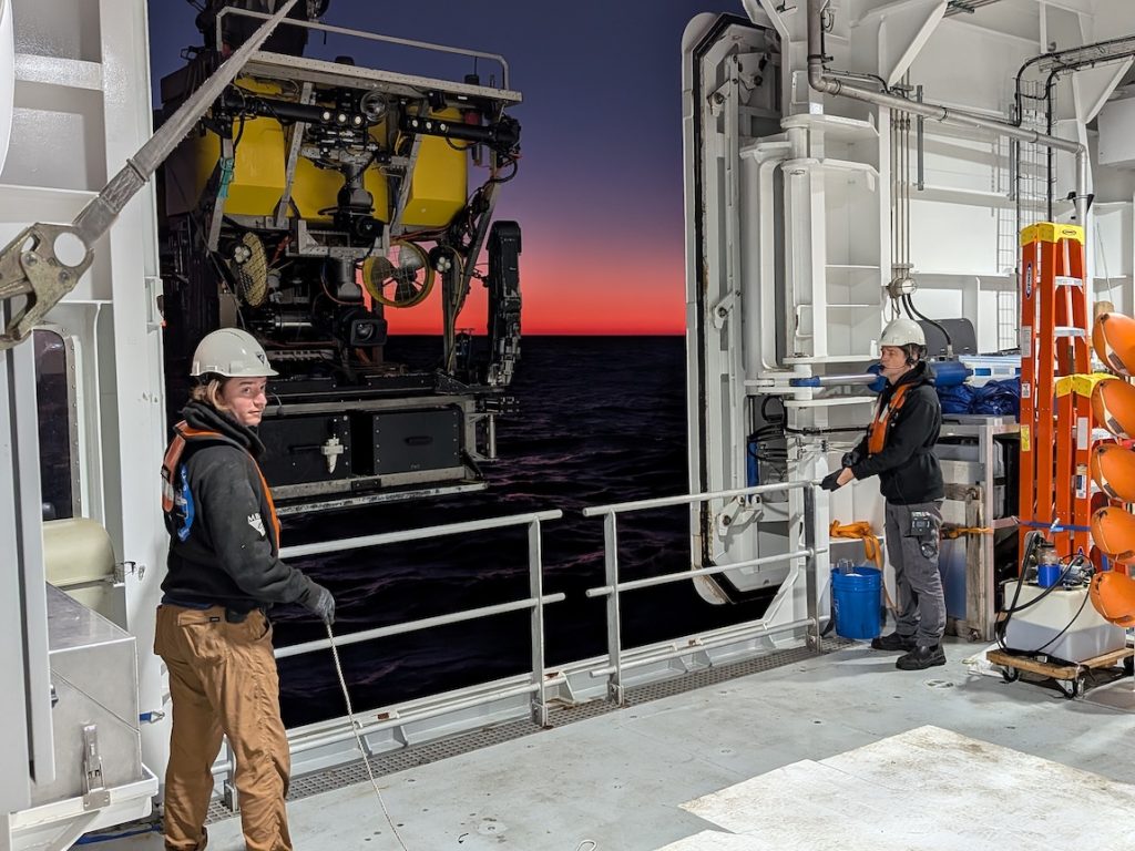 Two submersible pilots tend to ropes while a crane out of frame deploys a robotic submersible. The pilot on the left is wearing a white hard hat, black headset, black sweatshirt, orange life vest, and brown pants. The pilot on the right is wearing a white hard hat, black headset, black sweatshirt, orange life vest, gray pants, and black shoes. They are standing inside the hangar on a research ship with open doors. Inside the hangar is assorted maritime equipment. Outside the hangar is a robotic submersible with a yellow float, silver metal frame, and black metal instruments, and a black plastic trunk. In the background is the ocean’s surface at sunrise with black water, dark blue sky, and red sunlight on the horizon.