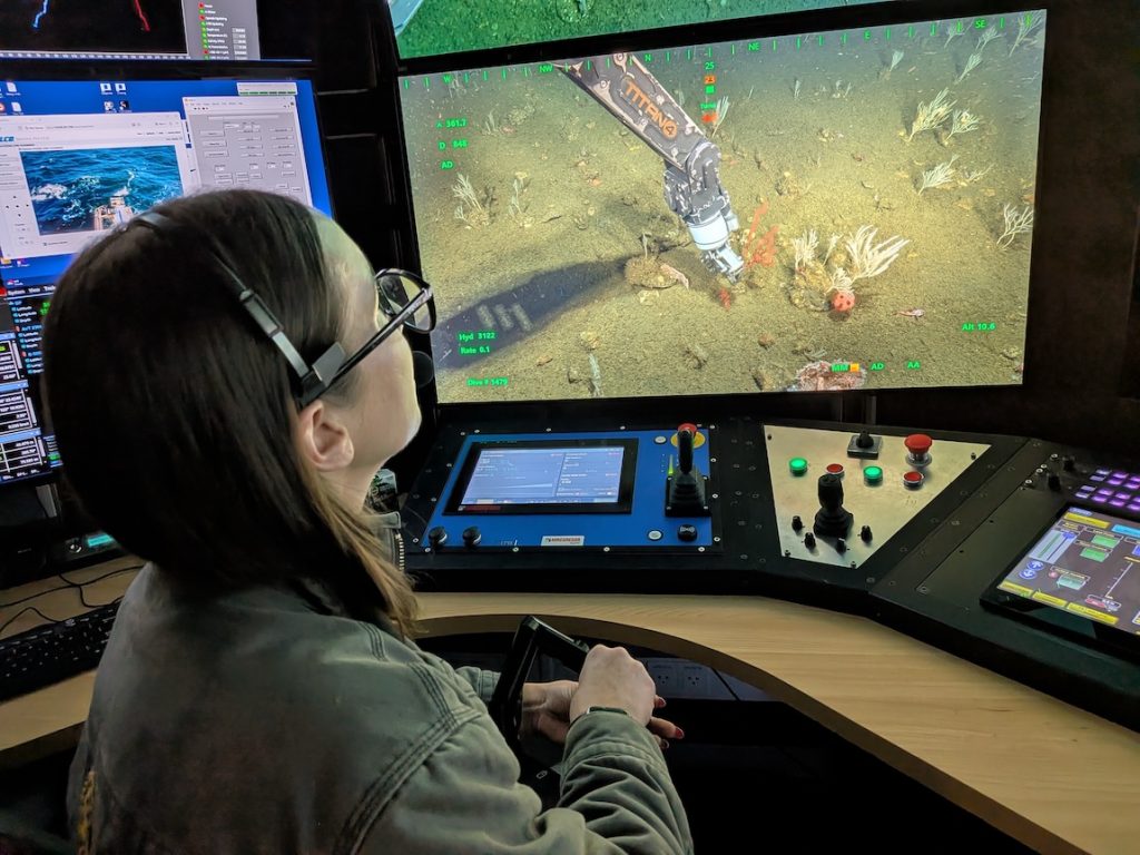 A submersible pilot operates the robotic arm of a deep-sea submersible from the dark control room of a research ship. The pilot has long brown hair and is wearing glasses, a black headset, and a green jacket. She is seated at a desk with various controls on the counter. She is holding a jointed black metal arm and watching a video monitor displaying the silver metal robotic arm of a submersible as it grips a rock on the deep seafloor attached to a red coral.