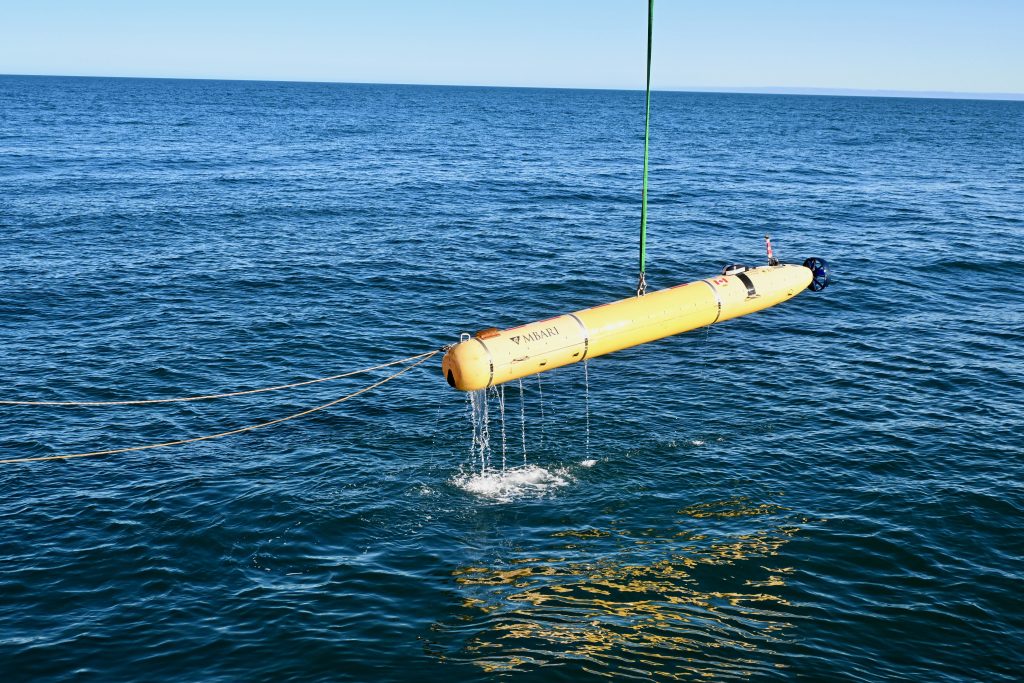 A large yellow, torpedo-shaped vehicle hangs from a line above the ocean. Two lines attach to the front of the vehicle and extend toward to the left of the image. The ocean is deep blue with small waves. The sky is light blue at the top of the image.