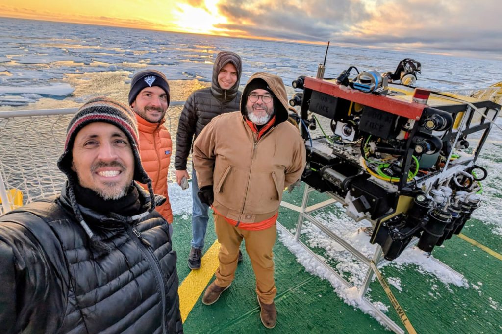 Four people in coats and hats stand on a green surface looking at the camera. They stand next to a remotely operated vehicle. The vehicle sits on a grey metal stand. A white rail with white netting is behind the people. In the background, the ocean is calm with a high density of sea ice. The bright yellow setting sun is just above the horizon of sea and ice. The sky is yellow and orange on the left side of the frame and filled with white and grey puffy clouds on the right.