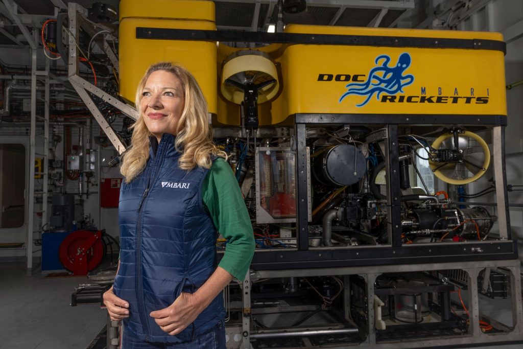 MBARI President and CEO Antje Boetius stands in front of a robotic submersible. She has shoulder-length blonde hair and is wearing a green shirt and blue vest with the MBARI logo. The submersible has a yellow float with a blue octopus logo and the words MBARI Doc Ricketts in blue and silver metal frame with assorted electronics and scientific equipment. In the background are assorted maritime equipment and the white metal walls of a hangar aboard an MBARI ship.