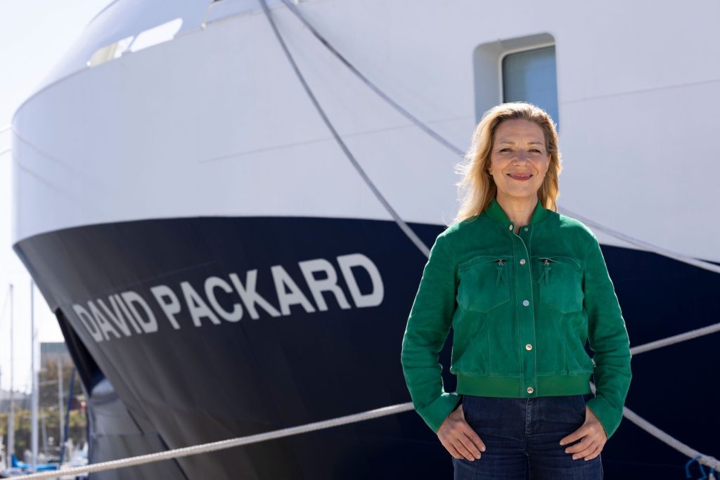 MBARI President and CEO Antje Boetius stands outdoors on a sunny day. She has shoulder-length blonde hair and is wearing a bright green jacket and dark blue jeans. In the background is a research ship with a blue-and-white hull and the name David Packard in white capital letters.