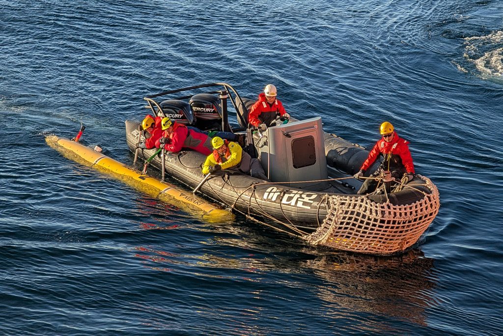 A small inflatable boat with two motors on the back holds five people with hard hats and all-weather gear. One person is steering the boat, one is sitting near the bow, and three are on the right side, holding a pole that attaches to a long, yellow, torpedo-shaped vehicle beside the boat. An MBARI logo is visible near the front of the vehicle. A white net covers the bow of the boat. The ocean surface is varying shades of blue to dark blue and has small ripples.