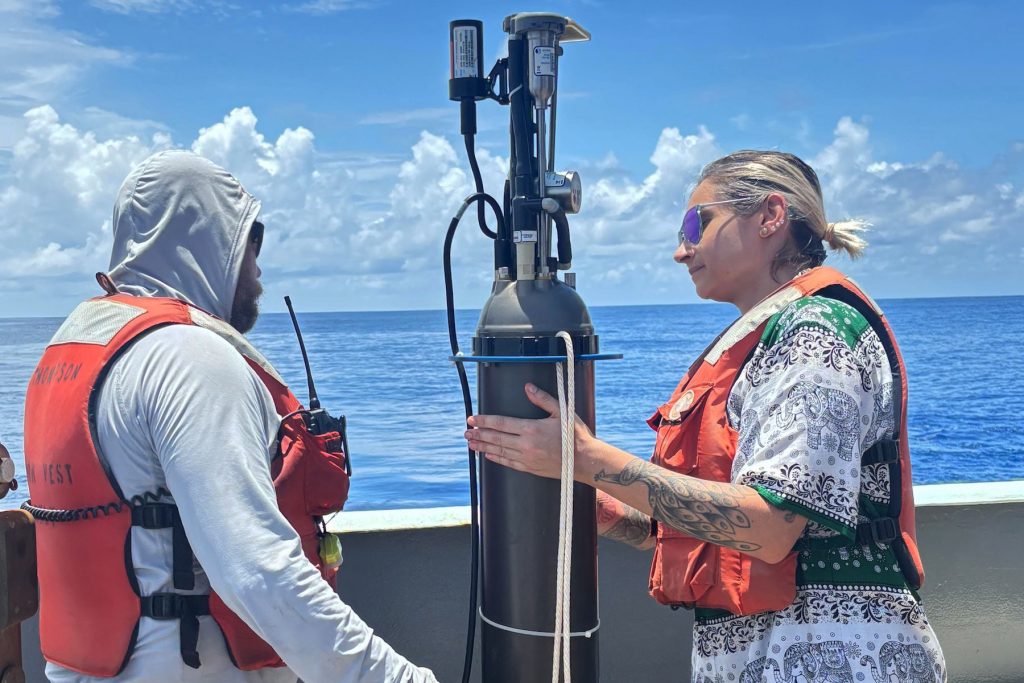 Two researchers prepare a robotic float for deployment. The researcher on the left has a brown beard and is wearing a white hooded sweatshirt and an orange life vest. The researcher on the right has blonde hair and is wearing sunglasses, a black-and-white patterned shirt, and an orange life vest. The cylindrical float has a gray plastic housing with black plastic instrumentation at the top. The researchers are standing on the deck of a research ship with a gray metal railing, blue ocean, blue sky, and white clouds in the background.