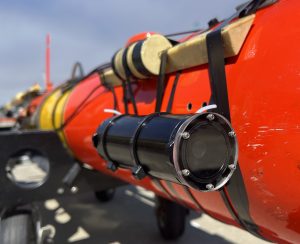 A black cylindrical camera is attached to pale yellow anchors with several strips of black plastic banding on the side of an orange-and-yellow underwater robot resting on a black plastic cart. This camera system was photographed outdoors on MBARI’s dock with hazy blue sky visible in the background.