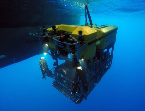 An MBARI remotely operated vehicle descends into the ocean. The robotic submersible has a bright yellow float on top of a black metal frame with a pair of black metal manipulator arms, a large camera, and several colored wires. The robot was photographed shortly after deployment, with its tether visible at the top of the frame. Bright blue water and one of the twin hulls of an MBARI research ship are visible in the background.